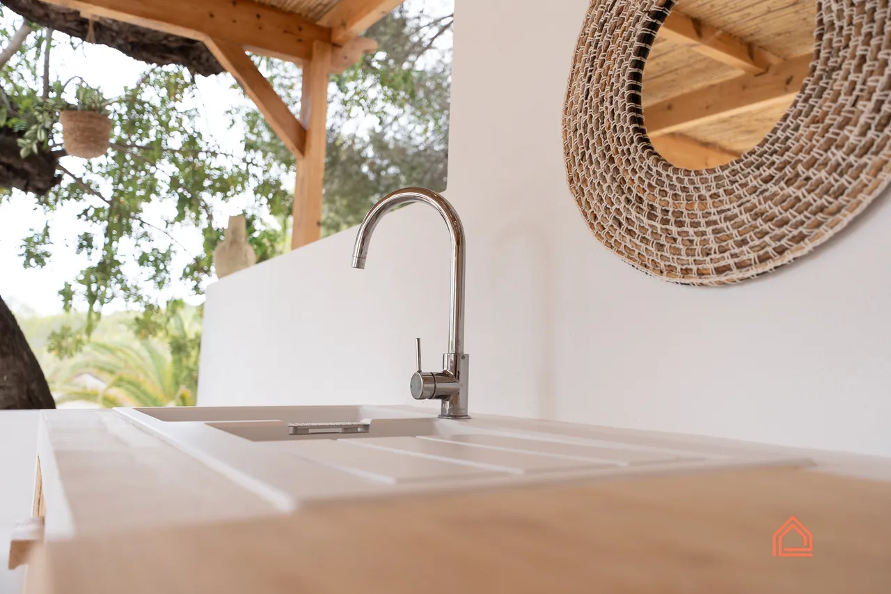 Outdoor kitchen sink with brushed steel tap and rattan mirror under a timber pergola